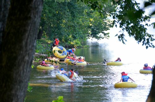 A group of people in colorful life jackets floating on yellow inner tubes in a calm river surrounded by trees.