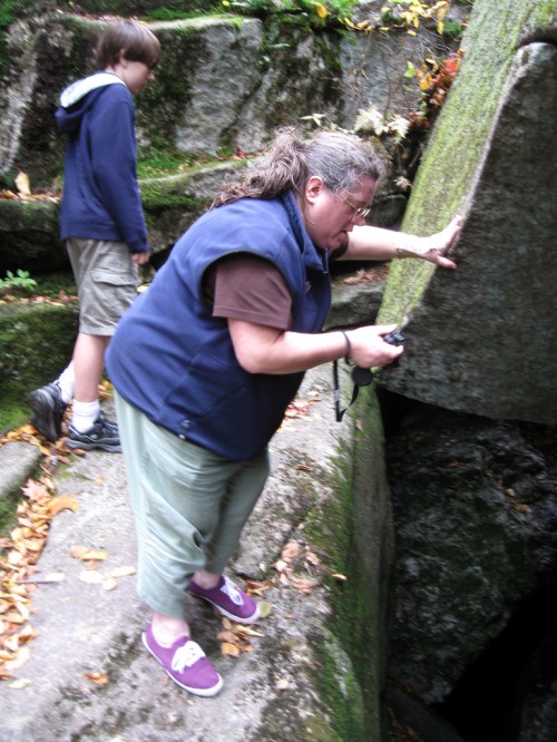 A woman takes a photo near a rocky area while a boy stands nearby, surrounded by moss and autumn leaves.