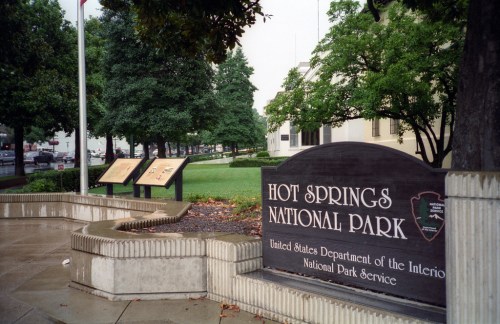 Sign for Hot Springs National Park with informational plaques and greenery in the background. Wet pavement reflects the scene.