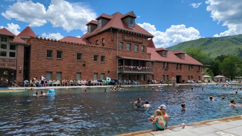 A historic building by a swimming pool with people enjoying the water on a sunny day, surrounded by mountains.
