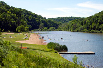 A serene lake surrounded by green trees, with people swimming and relaxing on the sandy shore.