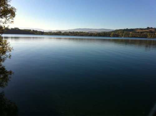 A calm lake reflecting the clear blue sky and surrounding greenery, with distant hills in the background.