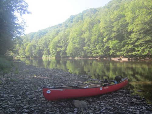 A red canoe rests on a rocky riverbank, surrounded by lush green trees and calm water reflecting the scenery.