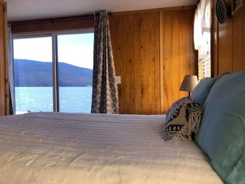 Cozy bedroom with a view of a lake and mountains, featuring a bed with blue pillows and wooden walls.