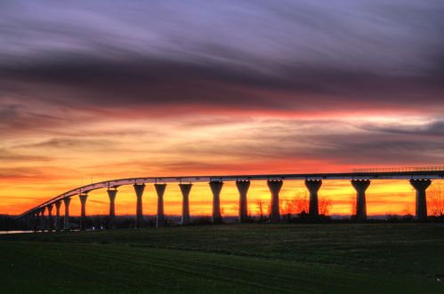 A bridge spans across a landscape at sunset, with vibrant colors in the sky and silhouettes of pillars.