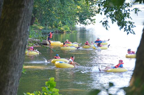 A group of people in yellow inner tubes floating on a calm river, surrounded by trees and greenery.