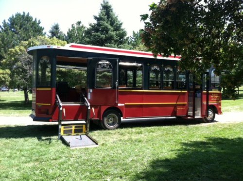 A red and black trolley bus parked on a grassy area, featuring a wheelchair ramp and surrounded by trees.