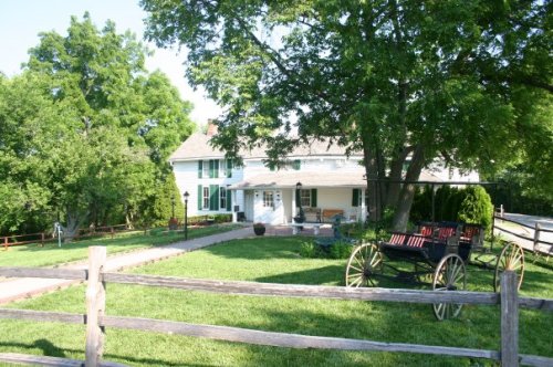 A charming white house with green shutters, surrounded by trees and a grassy yard, featuring a vintage horse-drawn carriage.
