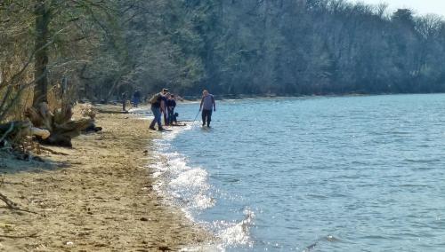 People walking along a sandy beach by a calm river, with trees in the background and gentle waves lapping at the shore.