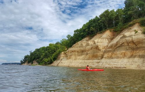A person in a red kayak paddles along a calm river near steep, sandy cliffs and lush green trees under a cloudy sky.