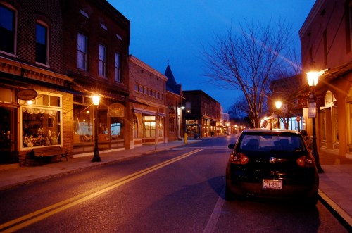 A quiet street at dusk, lined with shops and streetlights, with a parked car on the side.