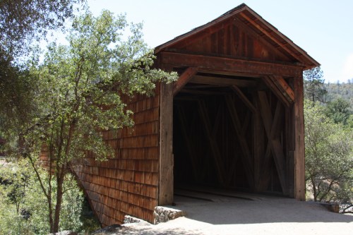 A rustic wooden covered bridge surrounded by trees, with a clear blue sky in the background.