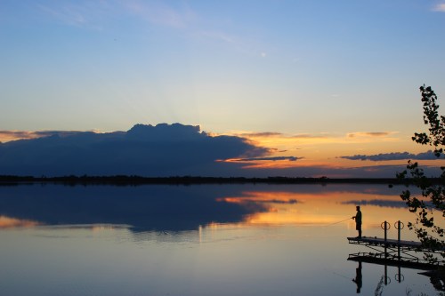 A serene sunset over a calm lake, with a lone fisherman on a dock silhouetted against the colorful sky.
