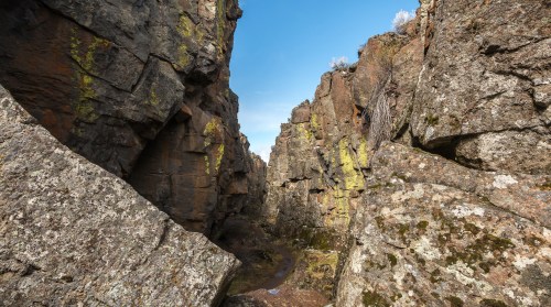 A narrow canyon surrounded by tall, rugged rock formations and patches of green moss under a clear blue sky.