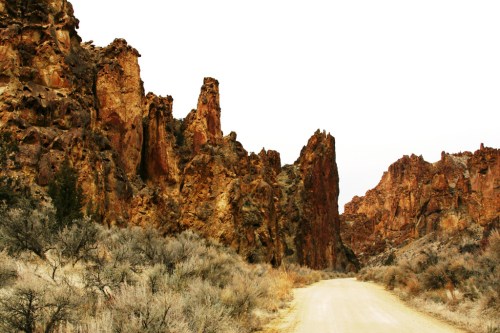 A dirt road winds through rugged, rocky terrain with towering cliffs and sparse vegetation under a cloudy sky.