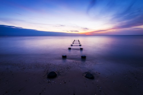 A serene beach at sunset, featuring a row of wooden posts leading into calm waters under a colorful sky.
