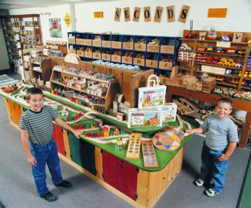 Two boys smile while playing with a colorful wooden train set and toys in a bright, organized playroom.