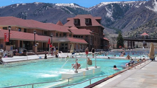 A sunny pool scene with people swimming, surrounded by mountains and buildings, featuring a clear blue sky.