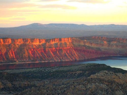 Sunset over a rugged landscape with colorful cliffs and a calm lake, surrounded by distant mountains.