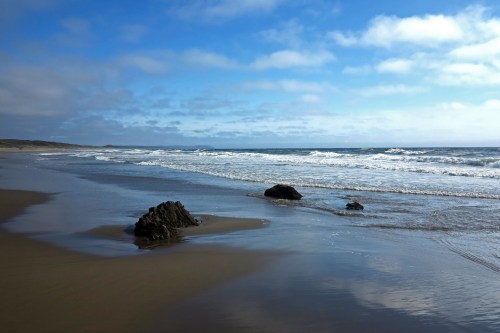 A serene beach scene with gentle waves, rocky formations, and a cloudy blue sky.