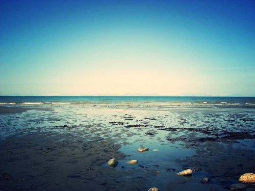 A serene beach scene with calm waters, distant mountains, and scattered rocks on the shore under a clear blue sky.
