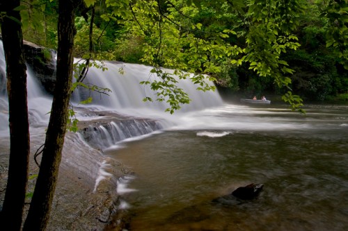 A serene waterfall cascades into a calm river, surrounded by lush green trees, with a small boat in the distance.