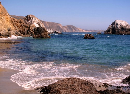 A serene beach scene with rocky cliffs, clear blue water, and gentle waves lapping at the shore under a clear sky.