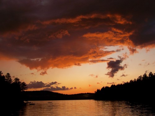 A serene lake at sunset, with vibrant orange clouds reflecting on the water and silhouettes of trees in the foreground.