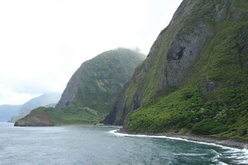 Lush green mountains rise steeply from a calm ocean, with a cloudy sky above and gentle waves lapping at the shore.