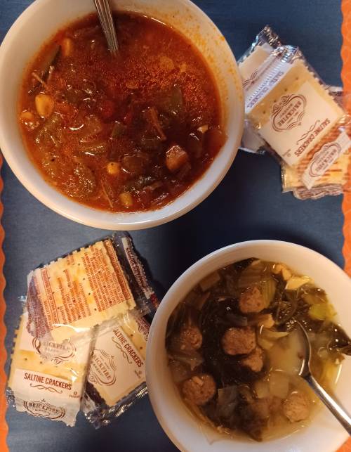 Two bowls of soup, one with meatballs and greens, the other with vegetables, accompanied by saltine crackers.