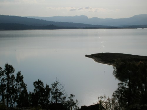 A serene lake reflects mountains and trees under a cloudy sky, with a small figure standing on the shore.