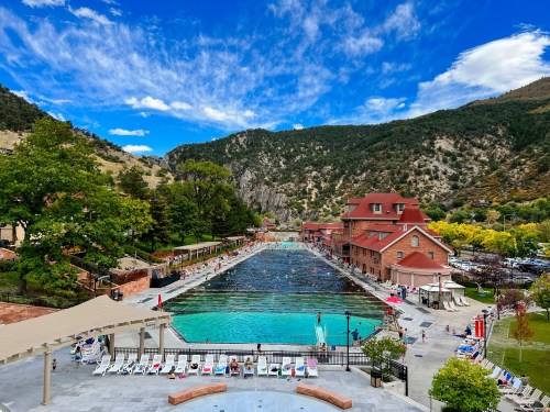 A scenic view of a large outdoor pool surrounded by mountains and trees, with people swimming and lounging nearby.