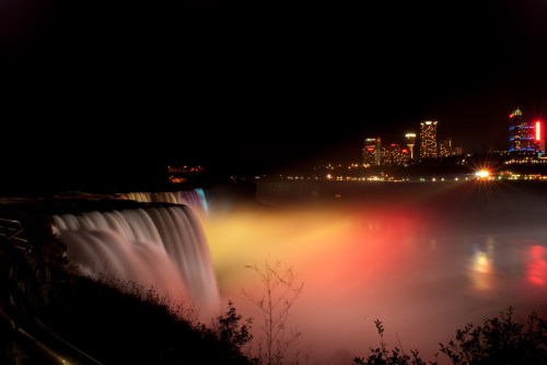 A nighttime view of Niagara Falls illuminated with colorful lights, with a city skyline in the background.