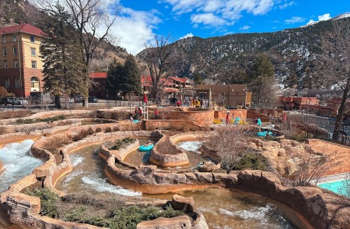 A scenic view of a winding water park with slides, surrounded by mountains and buildings under a blue sky.