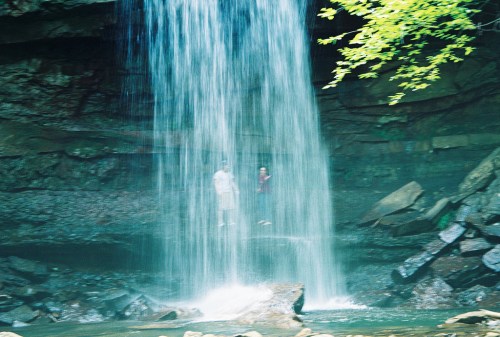 Two people stand behind a waterfall, surrounded by rocks and greenery, with water cascading down in front of them.