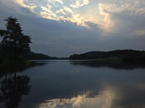 A serene lake reflects a cloudy sky, surrounded by trees and hills in the distance.