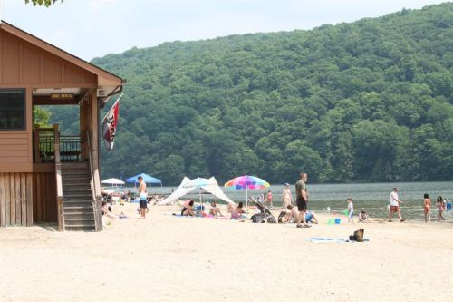 A sandy beach with people enjoying the sun, colorful umbrellas, and a wooden building near a lake surrounded by green hills.