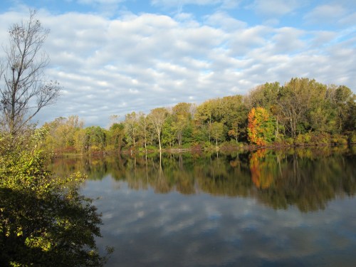 A serene lake surrounded by trees, reflecting colorful foliage and a cloudy sky.
