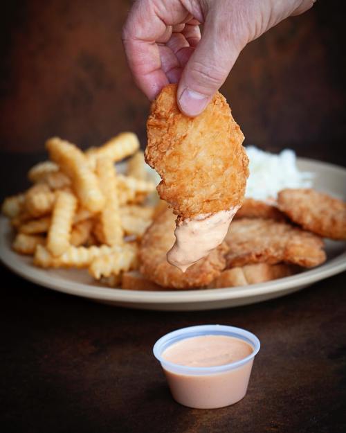 A hand dips a crispy chicken tender into a small cup of sauce, with fries and coleslaw on a plate in the background.