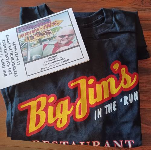 A folded black t-shirt with "Big Jim's" in red and yellow, next to a menu featuring a smiling man.