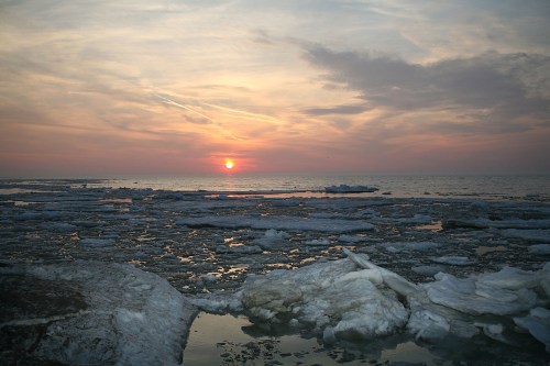 Sunset over a frozen lake, with ice formations and a colorful sky reflecting on the water's surface.