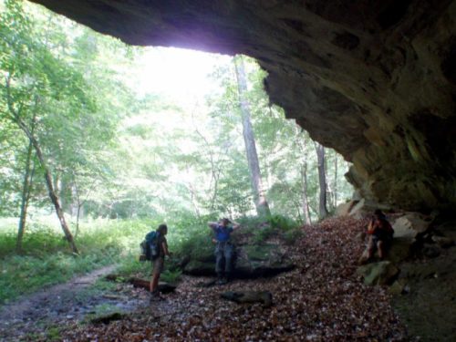 Two hikers explore a cave entrance surrounded by lush greenery and fallen leaves. Sunlight filters through the trees.