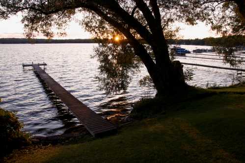 A serene lakeside scene at sunset, featuring a wooden dock extending into the water and trees framing the view.