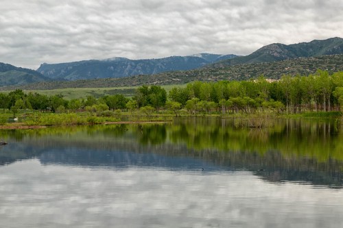 A serene lake reflects lush greenery and mountains under a cloudy sky.