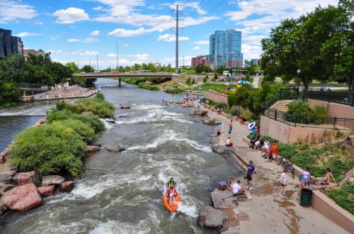 A lively river scene with kayakers, people enjoying the shore, and city buildings in the background under a blue sky.