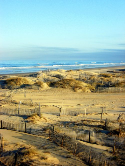 A serene beach scene with sand dunes, wooden fences, and gentle waves under a clear blue sky.