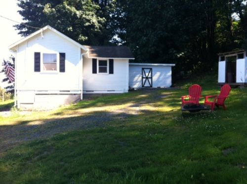A small white house with a gray roof, surrounded by green grass and two red chairs in front of a fire pit.