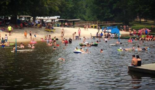 A crowded beach scene with people swimming and relaxing in the water, surrounded by trees and recreational areas.
