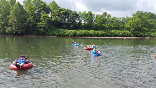 People floating on inner tubes in a calm river surrounded by lush greenery and a cloudy sky.