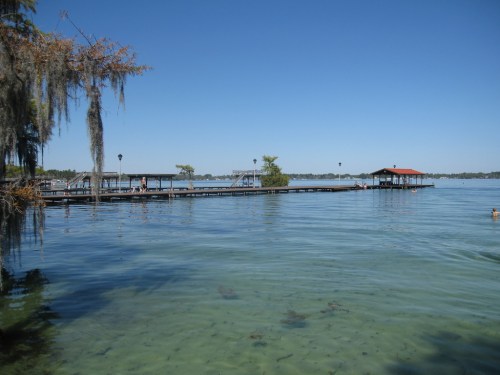 A serene lake view with a wooden dock, clear water, and a small boathouse under a clear blue sky.